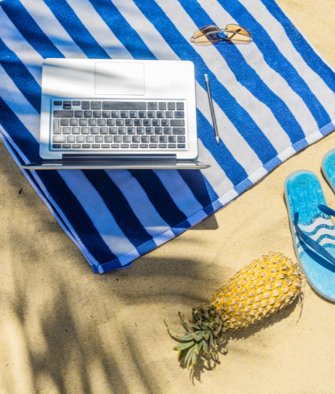 Imagen de una toalla sobre la arena de la playa con un portátil sobre ella, unas chanclas, una piña y unas gafas de sol